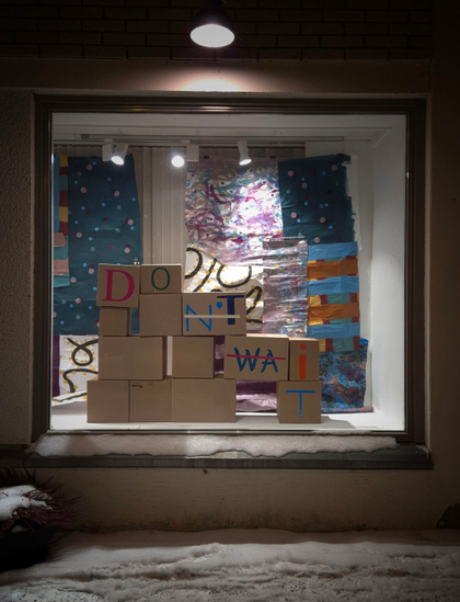 An art installation in a shop window. Several brown cardboard boxes are stacked on top of each other. The letters of the words ‘Don't wait’ are written across the boxes, with the letters ‘n't’ and “wa” crossed out so that it can also be read as ‘Do it’