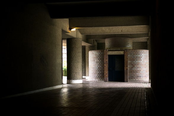 Photo looking along a dark space that ends at a circular dull garnet brick structure with a black wire mesh door between two openings to the outside world. The brick floor uses the same material as the structure and on the left there's at least one round brut concrete pillar.