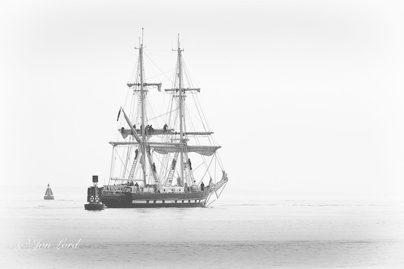 This is a low contrast black and white photo in landscape format of a square rigged sailing ship underway and about to vanish into a bank of sea fog. Southampton Water, UK (2019).

The lower quarter of the photo is filled with calm, flat and light grey sea water. There is an ill defined horizon with a thin sea fog, (a visibility of about one km), that obscures the sky and anything beyond. Very slightly to the right of centre is a two masted, tall ship, a brig, underway from left to right. The vessel has a historic look with a black hull, a thick white stripe running the length of the hull with square windows set in the stripe giving it the appearance of gun ports. Above the deck is a white deckhouse with 3 portholes and is located between the two masts. Stretching from the sides of the deck upwards to the first yard arm are ratlines, rope ladders, with sailors dressed in black, climbing up. Each mast has 3 yard arms with 6 sailors on the middle yard of the aft mast unfurling the sail. There are 2 further sets of ratlines, 1 reaching the upper yard, the other stretching to the mast head. The ship has just passed between 2 large navigation buoys marking the deep water channel. The nearer buoy has the words 'Black Jack' in bold white letters. 

Our ship is the 34m, 'TS Royalist'. A training ship, hence 'TS', owned and operated by the The Marine Society and Sea Cadets, carries 536 sq m of sail, has a permanent crew of 8, plus up to 24 Cadets and 2 adult trainees. 