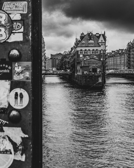 Aufnahme von dem Wasserschloss in Hamburg von der Poggenmühlen-Brücke aus gesehen. Dunkle Wolken schieben sich von rechts in das Bild hinein. Links im Foto sieht man noch einen Teil von einem genieteten Stützpfeiler der Brücke der mit diversen Aufklebern beklebt wurde. Das Foto ist in Schwarzweiß. 