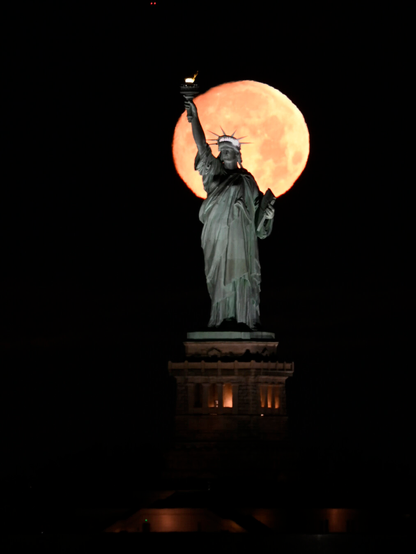 A full moon sets behind the Statue of Liberty. Her torch extends just beyond the perimeter of the moon's circumference.