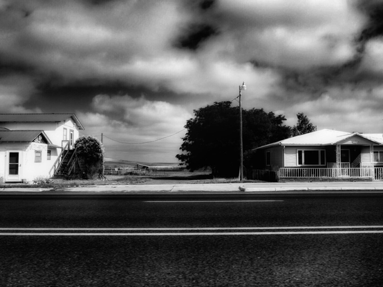 A black and white photo showing a wide angle view of two small houses by the side of a highway with a dramatically cloudy sky above.