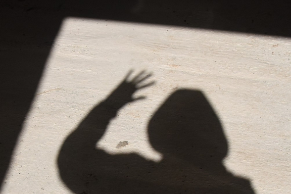 Shadow of a person the floor. Visible are a hooded head, one shoulder, one arm and one hand with fingers spread. The shadow is caught in a rectangle of sunlight surrounded by very dark shadow. 