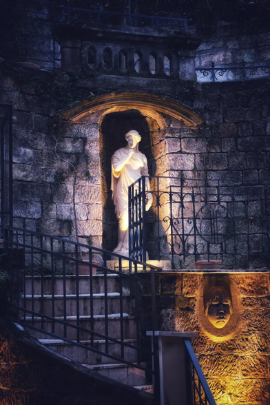 A nighttime scene of a stone statue illuminated in a recessed archway. The statue, resembling a classical figure, stands on a small balcony with a wrought-iron railing, set into a dark, textured stone wall. Warm lighting highlights the statue and casts a glow on the surrounding stonework, creating a dramatic contrast with the shadows. Below, a set of stone steps with a metal handrail leads up to the balcony. To the right, a carved stone face is subtly lit, adding to the mysterious and atmospheric ambiance of the setting.