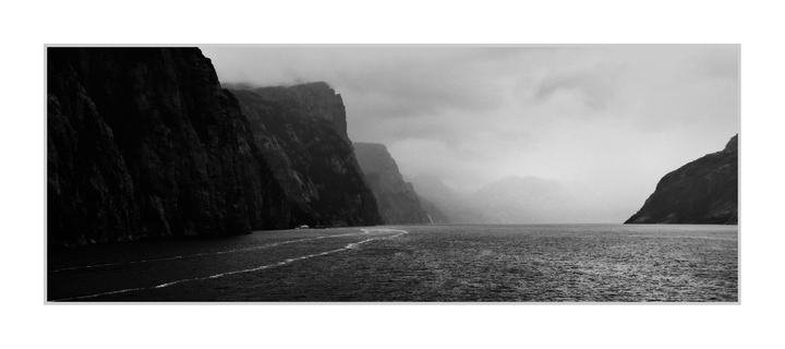 Wide panoramic black-and-white photograph. A broad expanse of dark water fills the lower half, textured by small ripples and crossed by thin, pale wake lines that angle toward the center distance. On the left, a steep, near-black rock wall dominates the frame and recedes into successive, lighter cliff faces fading into mist. On the right, a smaller dark headland slopes into the water, leaving a wide opening toward a bright, hazy horizon where the far shoreline and mountains are barely visible through low cloud.