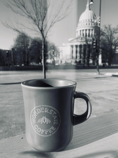 Black and white photo of a coffee mug on a ledge in front of a window. The Wisconsin state capitol building can be seen in the background. The coffee mug has the words “wonderstate coffee”.