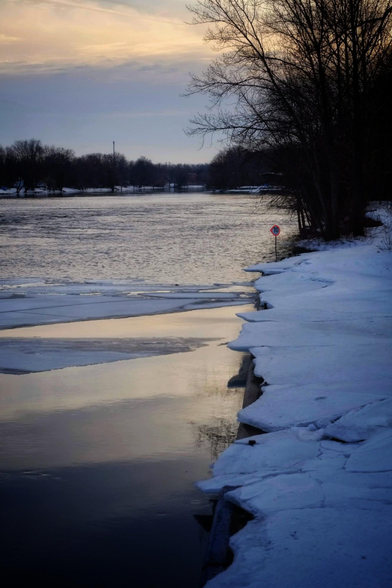 A river bank covered in snow and overhanging ice is seen at sunset. The wide river catches the colours of the early evening sky and large trees can be seen along the right side riverbank and in the distance.