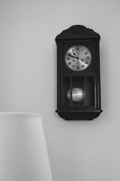 An einer weißen Wand hängt eine Pendeluhr, die 10:49 anzeigt. Das schwingende Pendel ist durch die Bewegung unscharf abgebildet. Links unten im Bild ist ein Lampenschirm ist ein Teil eines weißen Lampenschirms zu sehen – nicht ganz im Fokus.

A pendulum clock hangs on a white wall, showing 10:49. The swinging pendulum is blurred by its movement. At the bottom left of the picture, part of a white lampshade can be seen—slightly out of focus.
