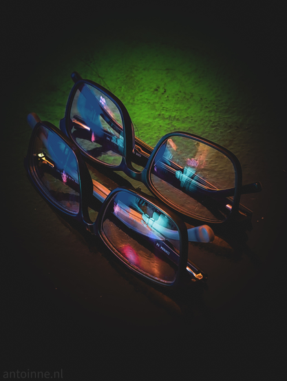 Two pairs of dark-rimmed glasses resting on a dark, textured surface. One pair is positioned slightly behind and above the other. 

The lenses act as mirrors, capturing distorted, colorful light patterns. A deep emerald green glow from above, with sharp highlights of magenta, cyan, and electric blue are reflecting off the lenses and frames.