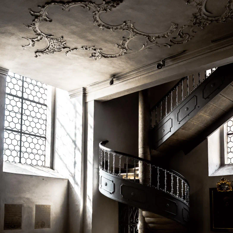 In der Ecke einer Kirche ist eine verzierte Wendeltreppe. Teile des Geländer bekommen Licht vom sonnendurchschienenen Fenster links daneben. Kontraste, farbgedämpft.
An der Decke Stuck.