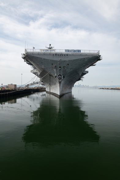 A symmetrical photograph of an aircraft carrier moored to a dock, head-on from the front, capturing its reflection in the water.