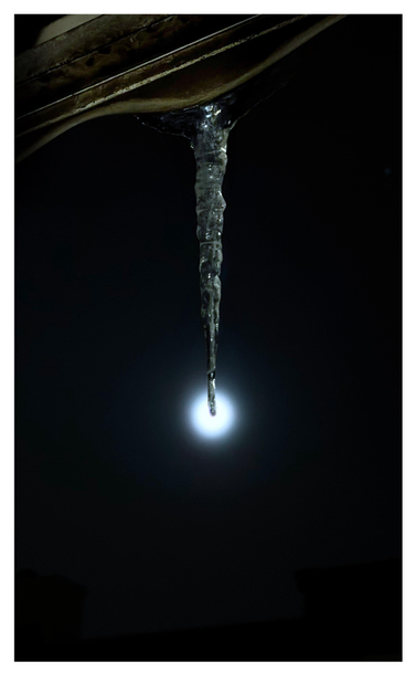 An evening in winter. A long icicle hangs down from the scalloped edge of a building awning. The icicle’s tip is perfectly aligned with the center of the full moon behind it, a small glowing white circle in a dark and featureless sky. The icicle and the underside of the awning are illuminated by a porch light behind the camera. The silhouettes of buildings at bottom frame can barely be seen, slightly darker shapes against the night sky.
