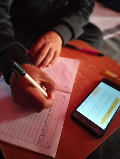 A close-up of a person's hand writing with a black pen on lined paper, with a smartphone displaying Arabic text on the right side of the frame. The hand holds the pen, moving it across the page. The smartphone rests on the table, its screen filled with text and a yellow button labeled in Arabic.