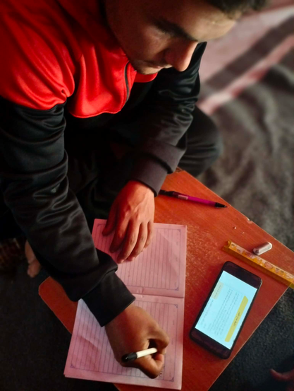 A person (Momen) wearing a black and red jacket is writing in a lined notebook on a small, wooden table, with a phone displaying text and other school supplies nearby.