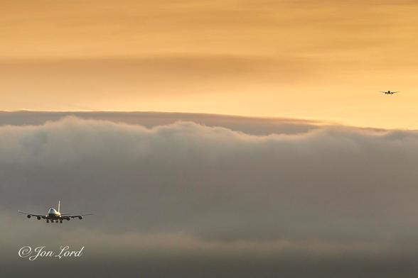 This is a colour sunrise photo in landscape format of two aircraft approaching to land. Los Angeles (2018).

The lower half of the image is filled with a layer of white, slightly fluffy, stratus cloud. The upper half of the photo has a near transparent layer of thin cirrus cloud filling the remains of the the photo, with darker stripes stretching across the frame from left to right and is rendered golden by the still hidden (behind the San Gabriel mountain range) rising sun. In the lower left corner is a large four engined passenger plane (Boeing747) with its wheels lowered on its approach to land. In the upper right corner is a further aircraft, possibly 5-10km behind the first. This jet has 2 engines and also has its wheels lowered. 
