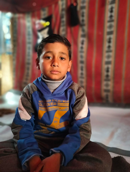 A young boy (Saji) with short dark hair sits cross-legged, looking directly at the camera. He wears a blue and gray hoodie with a light gray turtleneck and the words "NEW FASHION STYLISH WEAR COLLECTION WEAR URBAN" visible. The background is a blurred interior with red and white striped panels.
