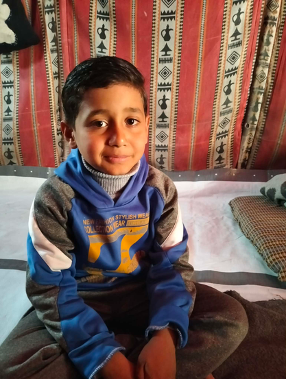 A young boy (Saji) with short dark hair sits on a bed, wearing a blue and gray hoodie with the words "New Fashion Stylish Wear Collection Urban" visible. He is looking at the camera with a slight smile, and behind him is a red, white, and black patterned tent wall.