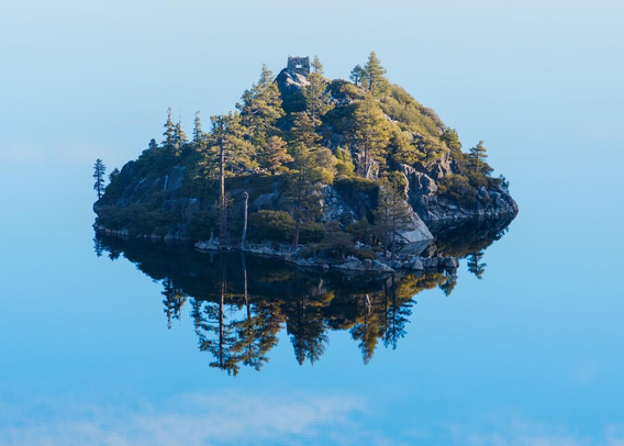 A color landscape photo of a small steep rocky island sitting in absolutely dead still blue water. It is early morning and the sun has just broke over an unseen ridge line to the right and is illuminating the top half of the island. The rocky island is pyramid shaped and cover in green bushes and conifer trees. There is a small abandoned stone structure at the top of the island. Because the water is dead still, the island is reflected, as if in a mirror, on on the water giving the effect that the island might be floating in the sky.