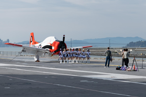 A row of “maid” cosplayers stand in front of a red and white military trainer aircraft on an aircraft carrier flight deck as a few photographers direct and shoot.