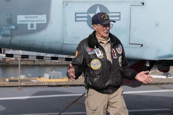 A man in a leather bomber jacket with patches from various vessel assignments, a baseball cap with “US Navy SCPO Retired” embroidered on it, and khaki uniform, stands before a vintage fighter plane on an aircraft carrier’s flight deck, speaking to a crowd off-camera.