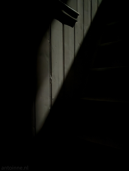 A sharp, diagonal beam of light cuts through the darkness, illuminating a section of a staircase and an adjacent light-colored wood paneled wall. 

The light reveals the physical details of the vertical wall panels, showing slight imperfections, grooves, and the matte finish of the paint. To the right, the edges of the wooden stairs are just barely visible, emerging from the gloom. The rest of the scene falls into deep, heavy shadow, leaving much of the environment to the imagination.