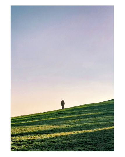 The photo is of a person walking down a grassy slope. The person is silhouetted against the sky. The hill is covered in bright green grass, with long shadows stretching across the surface. The sky above occupies a large portion of the photo, transitioning from a pale blue or light purple at the top to a warm, soft yellow near the horizon line where the hill meets the sky.