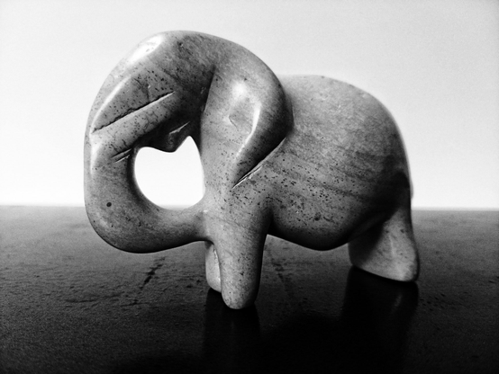 A black and white image of a smooth carved stone elephant standing on a dark table against a pure white background.