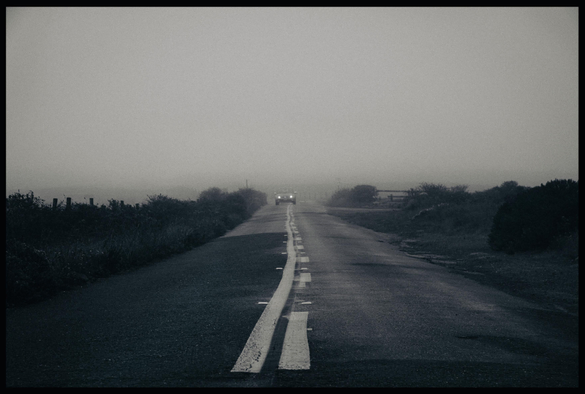 A dark scene on a very foggy morning at Point Reyes National Park. It's a picture of a small passenger car, with headlights on, driving in the middle of an asphalt, two lane road. The car somewhat straddles the lane marker in the center of the road, carefully avoiding the road edges because you never know when a deadly animal might leap out and attack. 

Okay I made that last part up. But what I didn't make up is the car is coming out of a heavy bank of fog and heading straight for me. Also, there are no trees... just small native bushes and weeds on either side of the road - the typical chaparral of this portion of the California coast.

Monochromey - slightly bluish and brownish dual tone but otherwise colorless - with limited dynamic range. There are no bright spots except for the car headlights. The sun would not come out for another 3 hours.