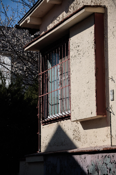 Close-up of a weathered window with a dull red metal grille, set in a textured beige wall, casting a shadow on the surface below.