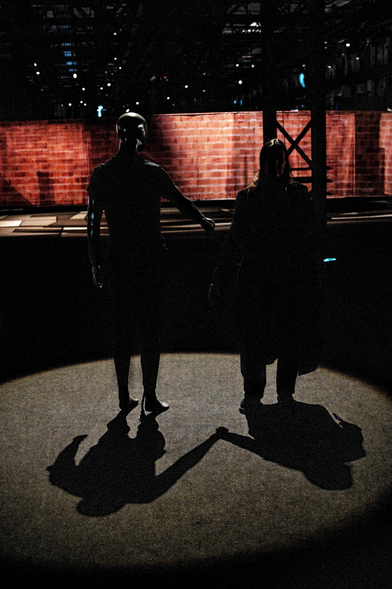 In einem runden Lichtkegel stehen eine Schaufensterpuppe und eine Person. Sie halten sich nicht an den Händen. Doch ihre Schatten auf dem Boden tun es.