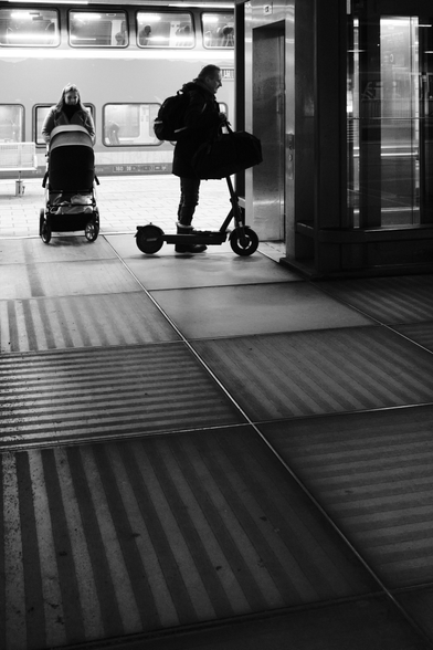 Ein Mann mit einem e-scooter und eine Frau mit einem Kinderwagen warten auf einem Bahnsteig vor einem Aufzug. Vor dem helleren Hintergrund werden sie teilweise als Silhouetten abgebildet. Sie stehen auf quadratischen gestreiften Bodenelementen aus Glas, die den Vordergrund des Schwarzweißfotos ausfüllen.. 

A man with an e-scooter and a woman with a stroller are waiting on a train platform in front of an elevator. Against the lighter background, they are partially depicted as silhouettes. They are standing on square striped glass floor elements that fill the foreground of the black-and-white photo.