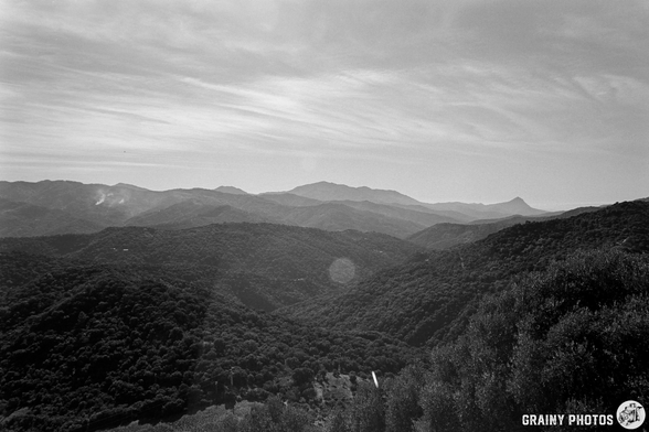 A black and white landscape featuring rolling mountains and lush forests, under a cloudy sky. The view captures a serene natural scene with varying elevations and distant peaks.