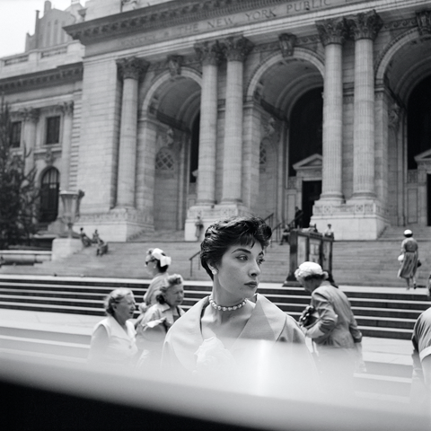 [Black-and-white photograph] In front of the colonnade entrance to the New York Public Library, a young, elegantly dressed woman is looking away from the camera at the presumably busy street.