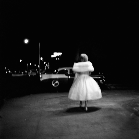 [Black-and-white photograph] A woman in a white dress and a pagola is walking away from the camera on the pavement at night. Her dress almost glows against the dark background.