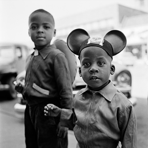 [Black-and-white photograph] Two boys, one wearing a Mickey Mouse ears hat, stand together on the pavement, looking curiously at the camera.