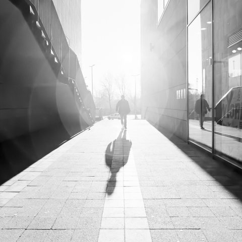 Ein Gang zwischen einer Treppe und einem Schaufenster. Gegen die Sonne eine Entgegenkommender, der sich auch im Schaufenster spiegelt. Quadratisch, s/w.
Hell.