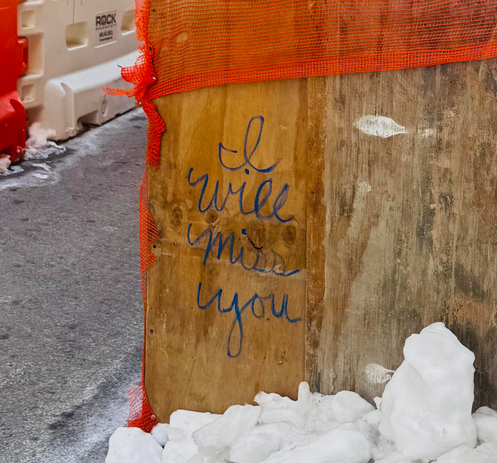 A color photo of the side of a construction site covered in old wooden boards. The right side of the photo is a plain wooden board and the left side of the photo show shows the street and some white and orange bollards. The middle of the photo is a wooden board and someone has written in script with a black marker enlarge letter letters "I will miss you" It made me a little sad. 