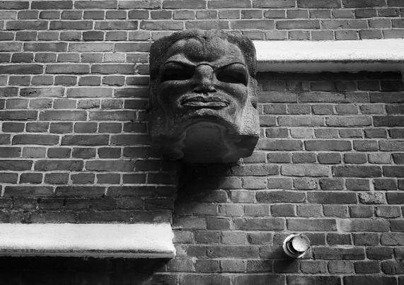 A dark stone sculpted face is mounted on a brick wall with two small white ledges below left and above right.
