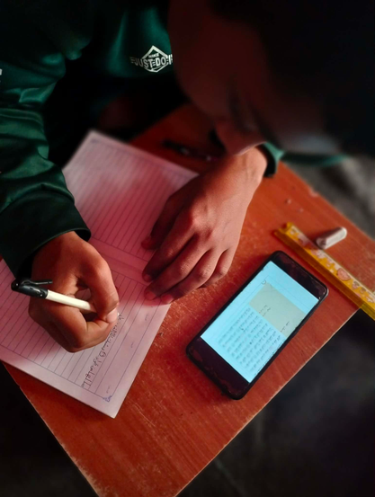 Abdul Rahman writing on a notebook with a pen on a wooden desk, with a smartphone displaying text and a ruler nearby. The person is wearing a green jacket.