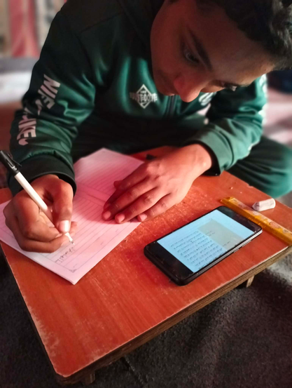 Abdul Rahman in a green sweatshirt writing on lined paper with a pen, a smartphone with text on the screen, a ruler, and an eraser are on a wooden desk.