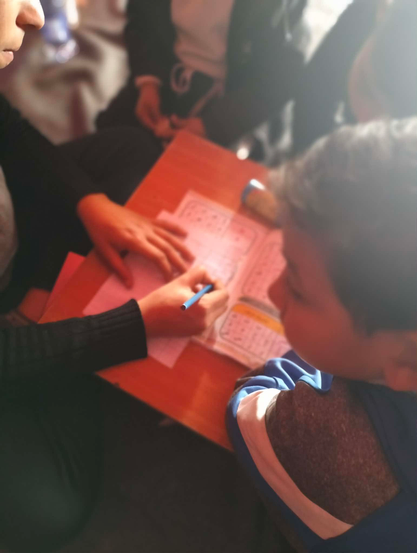 A person, writing with a blue pen on a sheet of paper with columns of printed numbers and symbols, on a wooden desk with other people. Another person is looking at the paper and person writing.