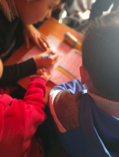 Children are writing on pink paper at a table, viewed from above, with one child's face partially visible, one child's arm in a pink sweater, and another child's head and neck in a blue sweatshirt facing away from the camera.