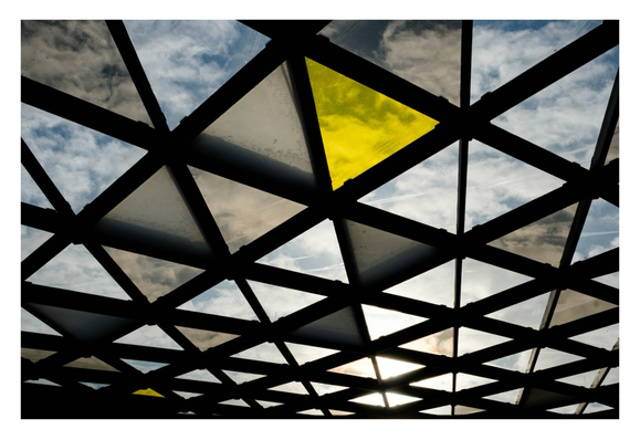 Foto im Querformat. Ausschnitt eines großen Glasdachs. Ein schwarzes Gitter bestehend aus Dreiecken. In den Dreiecken ist jeweils Glas, teils durchsichtig, teils gelb, teils grau. Darüber ist der bewölkte Himmel.