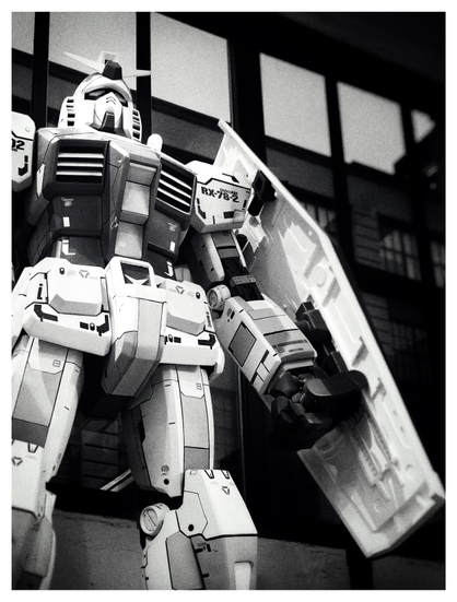 Black and white photo of a large model of a robot from the Gundam anime franchise, seen from the legs up, shot from below looking slightly upwards. The robot stands erect in a protective pose, its left (viewer’s right) fist clenched and carrying a long hexagonal shield. Its other arm is cropped from view. In the background are shadowy window panels with a view of buildings and the evening sky outside.