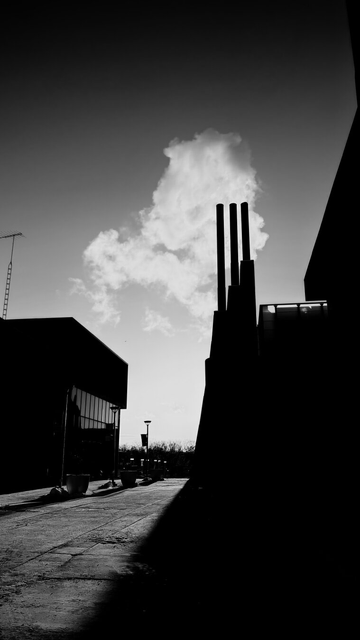 View in betwren 2 buildings. The one of the left is low, with a reflective line of windows. The one on the right is high, looming, in shadow, with 3 smokestacks. Low angled sunlight casts between the two buildings, illuminating the smoke coming from the stacks and the rough concrete between the buildings. Black and white photo.