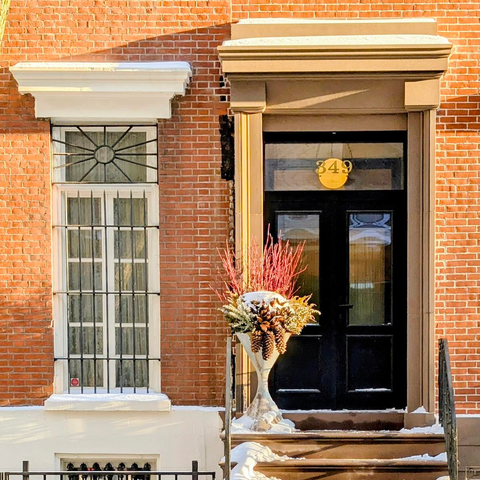 A bright red brick building featuring a sleek, modern black double door numbered 349. A large stone urn filled with winter greenery and red branches sits on the stoop. To the left, a white-trimmed window is protected by a decorative sunburst iron grate.