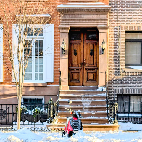 A wide shot of a classic Chelsea brownstone with a high stone stoop and ornate double wooden doors. A modern red and black electric scooter is parked at the bottom of the stairs, partially buried in a large snowbank on the sidewalk.
