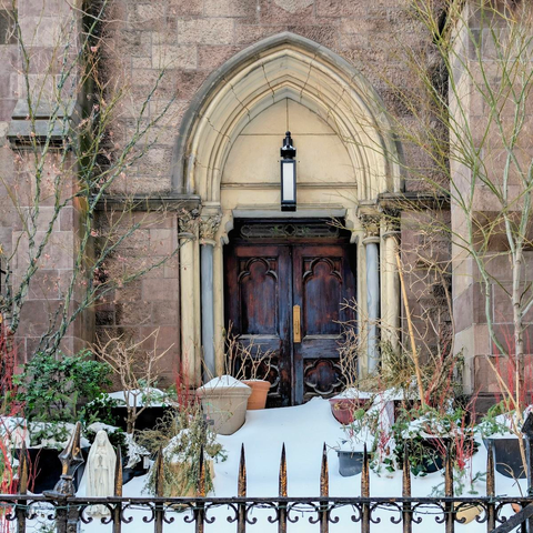 A dark, weathered wooden door with Gothic-style quatrefoil carvings set into a pointed stone archway. Deep snow piles up against the base of the door, surrounding various dormant potted plants and a small white statue of the Virgin Mary.