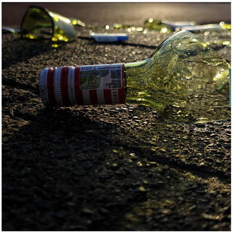 A photo showing the top of a broken glass bottle with a red and white striped label on the pavement. Pieces of greenish glass are visible in the background against low sunlight.