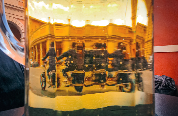 The photo is of a distorted reflection of a street scene with a person on a bike, as seen through a glass of beer.
The reflection has a grand, yellow building facade with a colonnade. On the street, blurred figures (it's just one person reflected a few times) on a bike are visible.The reflections are warped and fragmented due to the curvature and thickness of the glass.